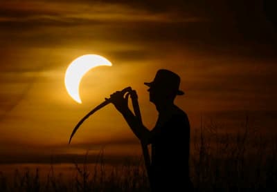 A farmer holding a scythe stands in a meadow before the solar eclipse in Jacobsdorf, Brandenburg, Germany. [Patrick Pleul/Picture Alliance via Getty Images]