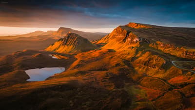 Golden light bathes the rugged highlands, where silent lakes mirror the dramatic peaks under a brooding, darkening sky. 🏔️