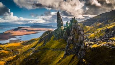 Ancient rocky spires stand tall against dramatic clouds, overlooking a serene blue loch and rolling golden highlands below. 🏔️